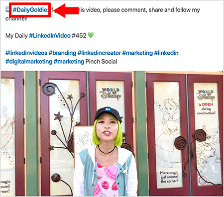 Dies ist ein Screenshot, der zeigt, wie Goldie Chan Hashtags im Text ihrer LinkedIn-Videobeiträge verwendet. Rote Beschriftungen verweisen auf das Hashtag #DailyGoldie im Text, das für ihre Videopostings eindeutig ist und ihr hilft, Freigaben zu verfolgen. Der Beitrag enthält auch andere relevante Hashtags, die den Nutzern helfen, ihr Video zu finden, einschließlich #LinkedInVideo. Im Videobild steht Goldie vor einigen Türen eines World of Disney-Displays. Sie ist eine asiatische Frau mit grünen Haaren. Sie trägt eine schwarze LinkedIn-Mütze, eine schwarze Chokerhalskette, ein rosa Hemd mit Macaron-Print und eine blau-weiße Jacke.