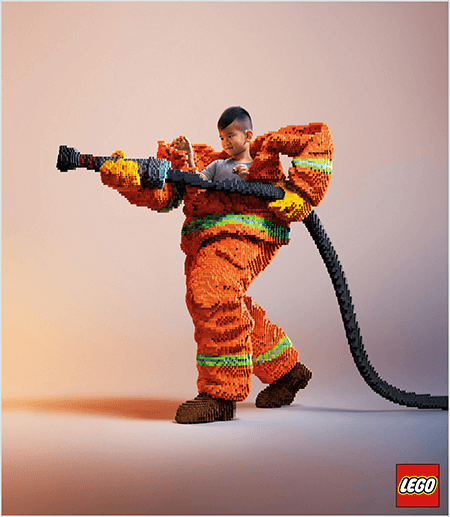 Dies ist ein Foto aus einer LEGO-Anzeige, das einen jungen asiatischen Jungen in einer Feuerwehruniform aus LEGOs zeigt. Die Uniform ist orange mit einem neongrünen Streifen um die Manschetten von Mantel und Hose. Der Feuerwehrmann steht mit einem Fuß zurück und hält einen Feuerwehrschlauch, ebenfalls aus Legos. Der Kopf des Jungen erscheint aus der Oberseite der Uniform, die viel größer ist als er, und bleibt um die Schultern stehen. Das Foto wurde vor einem einfachen neutralen Hintergrund aufgenommen. Das LEGO Logo erscheint in einem roten Feld unten rechts. Laut Talia Wolf ist LEGO ein großartiges Beispiel für eine Marke, die Emotionen in der Werbung einsetzt.